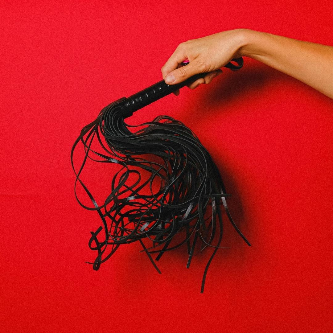 Black flogger held by a hand against a red background